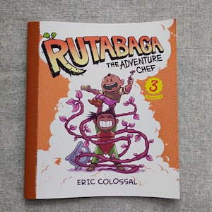 May include: A softcover graphic novel titled Rutabaga The Adventure Chef 3 by Eric Colossal. The cover features a cartoon illustration of two characters, one lifted above the other by purple vines, with a small cooking pot and orange background.