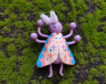 May include: A purple creature figurine with six limbs ending in spheres, two fuzzy white antennae, and two large wings. The wings are painted with a cream base, featuring blue and coral scalloped borders, and decorated with small purple and coral stars.