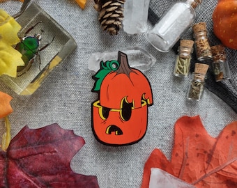 May include: A cartoon-style orange pumpkin with a carved face and a green stem. The pumpkin is the focal point, surrounded by fall-themed decorations like leaves, crystals, and small glass bottles with corks. The overall theme is Halloween.