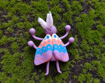 May include: A purple insect figurine featuring two white fuzzy antennae and four arms ending in spheres. The wings are painted with horizontal bands of orange, blue, and purple, decorated with small dots in contrasting colors throughout the design.