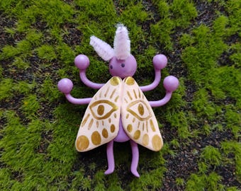 May include: A purple creature figurine with four arms ending in spheres, a round head with white fuzzy antennae, and two light pink wings featuring gold painted eye motifs and radiating lines. The body is smooth and solid in construction.