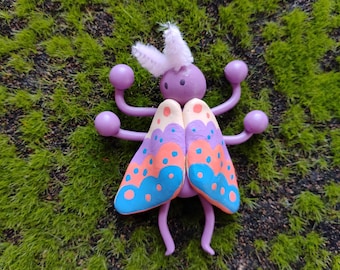 May include: A handcrafted insect figurine featuring a purple head with white fuzzy antennae, six purple limbs ending in spheres, and two wings painted with scalloped patterns in shades of orange, blue, and purple with small blue dots.