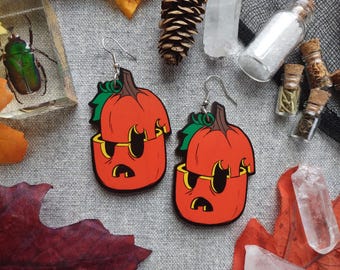 May include: A pair of orange pumpkin earrings with green leaves and a surprised face. The earrings have a black outline and are hanging from silver hooks. The background includes fall-themed decorations.