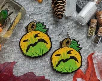 May include: A pair of Halloween-themed earrings featuring a pumpkin design. The earrings have a yellow and green color scheme with a black, sad face. The earrings are made of wood and have silver-colored hooks.