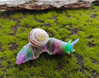 May include: A small snail figurine featuring a translucent, multi-colored body with swirls of pink, green, and blue. The snail has a realistic, spiraled, off-white shell with thin, concentric brown stripes positioned on its back.