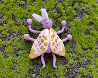 May include: A decorative insect figurine featuring a smooth lavender body with four curved limbs ending in small spheres. It has two fuzzy white antennae and two large, cream-colored wings adorned with metallic gold painted spots and patterns.