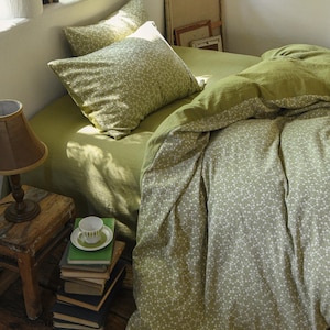 Könnte beinhalten: Ein Bett mit olivgrüner Bettwäsche und Kissen. Der Bettbezug und die Kissenbezüge weisen ein weißes Blumenmuster auf. Ein kleiner Holzschemel trägt einen Stapel Bücher und eine Teetasse. Eine Lampe mit braunem Lampenschirm steht auf dem Hocker.