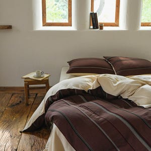 May include: A bedroom scene featuring a bed with a brown and cream striped duvet cover and matching pillows. The bed is against a white wall with wooden framed windows. A small wooden side table holds a cup and saucer.