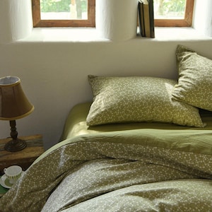 Può includere: Un letto con biancheria da letto verde oliva e pois bianchi. Sono visibili due cuscini abbinati. Una piccola lampada con paralume marrone è su un comodino di legno vicino a una tazza e un piattino.
