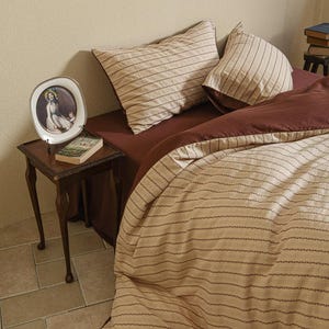 May include: A bed with a brown duvet cover and pillowcases. The bedding has a beige background with thin, dark brown horizontal stripes. A small side table holds a framed picture and a book. The bed is against a cream wall.