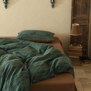 May include: A teal and brown striped duvet cover with a teal and brown striped pillow on a brown bed. A lamp with a brown lampshade is on a stack of books to the right of the bed.