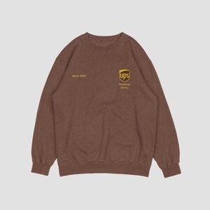 Peut inclure: Sweat-shirt marron avec le logo UPS et le texte "Worldwide Service". Le sweat-shirt comporte également le texte "since 1907". Le design est de couleur jaune. Le sweat-shirt est de couleur marron uni.
