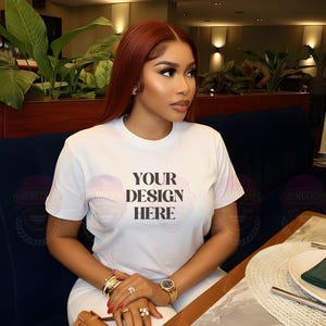 Könnte beinhalten: Weißes T-Shirt mit dem Text "YOUR DESIGN HERE". Die Person trägt Goldschmuck, darunter eine Uhr und Armbänder, und hält eine hellbraune Handtasche. Der Hintergrund umfasst eine dunkelblaue Sitzbank und einen Marmortisch.