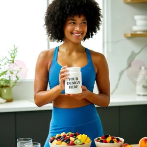 Puede incluir: Una mujer con ropa deportiva azul sostiene un tarro blanco con el texto "YOUR DESIGN HERE". Cuencos de fruta fresca, incluyendo bayas, kiwi y mango, están sobre una superficie de madera. La escena tiene lugar en una cocina luminosa.
