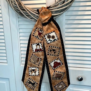 May include: A decorative scarf featuring a farm theme. The scarf has a tan background with black polka dot trim and square images of farm animals and objects. The scarf is hanging on a white door.