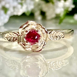 May include: A silver ring with a red gemstone set in a rose design. The band of the ring is decorated with leaves.