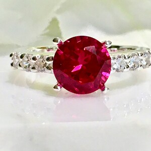 May include: A silver ring with a large, round, bright red gemstone surrounded by a band of smaller, clear gemstones.