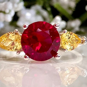 May include: A silver ring featuring a large, round, red gemstone flanked by two smaller, pear-shaped, yellow gemstones.