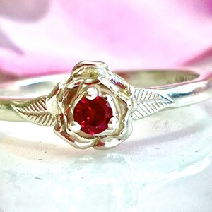 May include: A sterling silver ring featuring a rose design. The ring has a central, round, bright red gemstone. The rose is detailed with petals and leaves, and the band is smooth. The ring is a piece of jewelry.