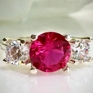 May include: A silver ring with a large, bright pink gemstone surrounded by two smaller, clear gemstones.