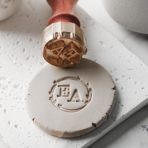 May include: A brass seal stamp with a wooden handle, pressed into a round, off-white clay impression. The stamp features the initials "EA" in a decorative font. The clay impression is on a white, textured surface.