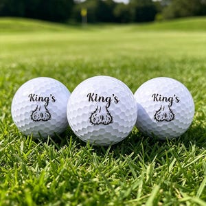 Könnte beinhalten: Drei weiße Golfbälle mit genoppter Oberfläche, jeweils mit dem schwarzen Aufdruck King's über einer schwarzen Strichzeichnung eines Hodensacks. Die Golfbälle sind in einer horizontalen Reihe angeordnet und zeigen das einheitliche Design.