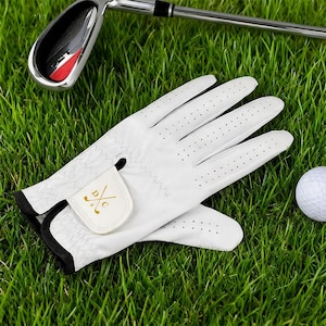 May include: White golf gloves with black trim and small perforations, resting on green grass next to a golf ball and a golf club. The gloves have a small patch with the initials "D C" and crossed golf clubs in gold.