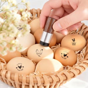 Könnte beinhalten: Ein Eierstempel mit Holzgriff wird verwendet, um frische Eier in einem Weidenkorb zu markieren. Die Eier sind mit den Worten "SIMON" und "Fresh eggs" sowie einer Hühnerzeichnung versehen. Die Eier sind hellbraun und beige.