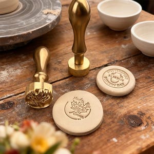 May include: A collection of pottery tools and supplies on a wooden surface. Includes two brass stamps, two small white bowls, and two round clay stamps with text and a maple leaf design. A pottery wheel is visible in the background.