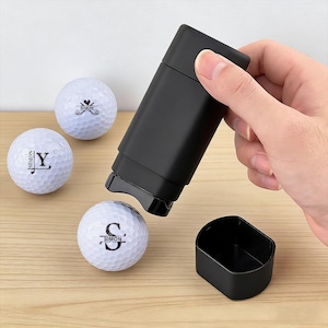 May include: A black golf ball stamper and three white golf balls with black personalized text and designs. The stamper is being used to mark a golf ball. The golf balls are inscribed with the names "Simon" and "JY".