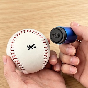 May include: A white baseball with red stitching and the black text MBC printed on the surface, accompanied by a cylindrical blue handheld stamping tool featuring a black circular end cap with the black text WBC embossed on the surface.