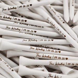May include: A pile of white wooden golf tees, each featuring the dark brown engraved text The Wades, the date 09 06 26, and a small heart symbol. The tees are arranged in a jumbled heap, showcasing the personalized branding on each piece.