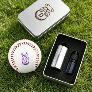 May include: A white baseball with red stitching and a purple bulldog graphic featuring the letter D, accompanied by a silver metal tin containing a silver cylindrical object and a small black bottle with a cap, both set in black foam.