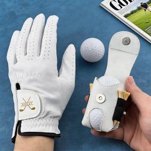Może przedstawiać: Biała rękawica golfowa z napisem "Ashley" i skrzyżowanymi kijami golfowymi. Biały skórzany uchwyt na piłeczki golfowe z dwiema piłeczkami i tee. Widoczna jest również biała piłeczka golfowa.