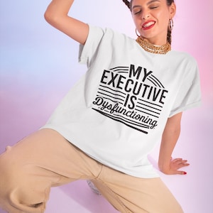 Puede incluir: Camiseta blanca con texto negro que dice "My Executive Is Dysfunctioning". El texto está en un diseño curvo y rayado. Una persona lleva la camiseta y tiene el pelo en una trenza.