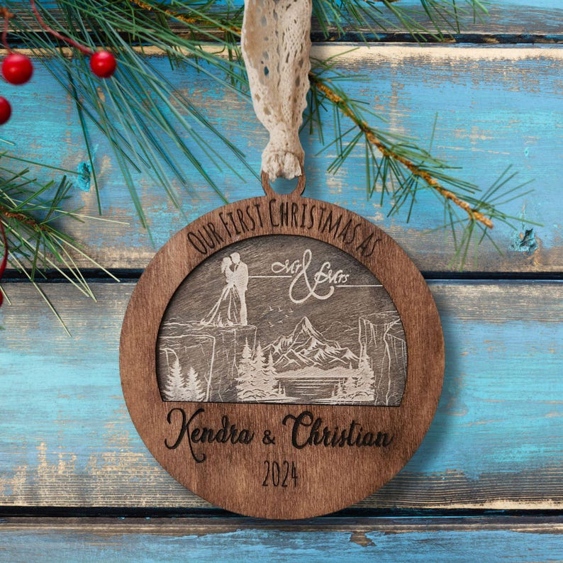May include: A round wooden Christmas ornament with a laser-engraved design. The ornament features a couple standing on a cliff overlooking a mountain range. The text "Our First Christmas as Mr. & Mrs." is engraved at the top of the ornament, and the names "Kendra & Christian" and the year "2024" are engraved at the bottom.