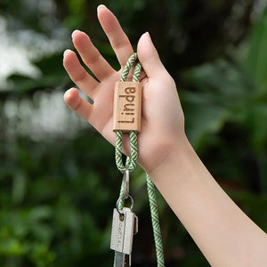Pode incluir: Um cord&atilde;o tran&ccedil;ado verde e castanho com uma etiqueta de madeira que diz "Linda" e um chaveiro prateado com uma chave.