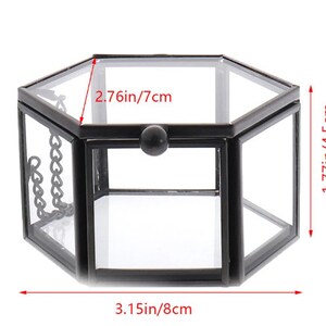 K&ouml;nnte beinhalten: Eine sechseckige Glas-Displaybox mit schwarzem Metallrahmen, Klappdeckel und kleinem Metallknopf. Die Box misst 8 cm in der Breite, 4,5 cm in der H&ouml;he und 7 cm in der Tiefe und verf&uuml;gt &uuml;ber eine kleine Kette an der Seite zur Deckelst&uuml;tze.