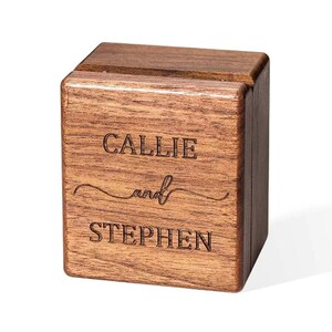 May include: A square, dark brown wooden ring box with a hinged lid. The front face features the laser-engraved text "CALLIE and STEPHEN" in a combination of block lettering and a cursive script font, showcasing the natural wood grain texture.