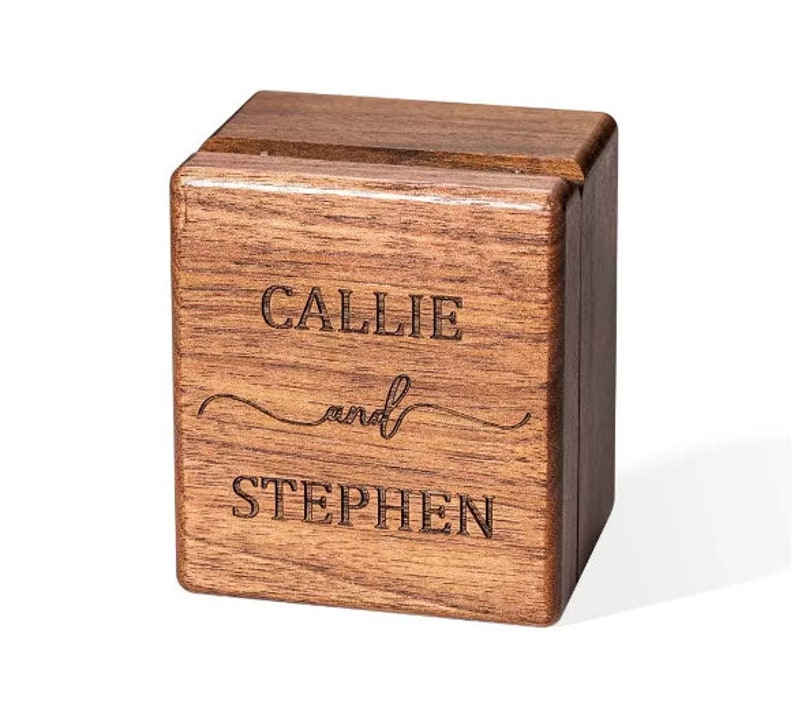 May include: A square, dark brown wooden ring box with a hinged lid. The front face features the laser-engraved text "CALLIE and STEPHEN" in a combination of block lettering and a cursive script font, showcasing the natural wood grain texture.