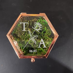 K&ouml;nnte beinhalten: Eine sechseckige Glasschatulle mit einem ros&eacute;goldenen Metallrahmen, gef&uuml;llt mit gr&uuml;nem Moos. Der Glasdeckel ist mit wei&szlig;em Text graviert, darunter die Buchstaben T und A, eine Strichzeichnung einer Rose und das Datum 06.03.