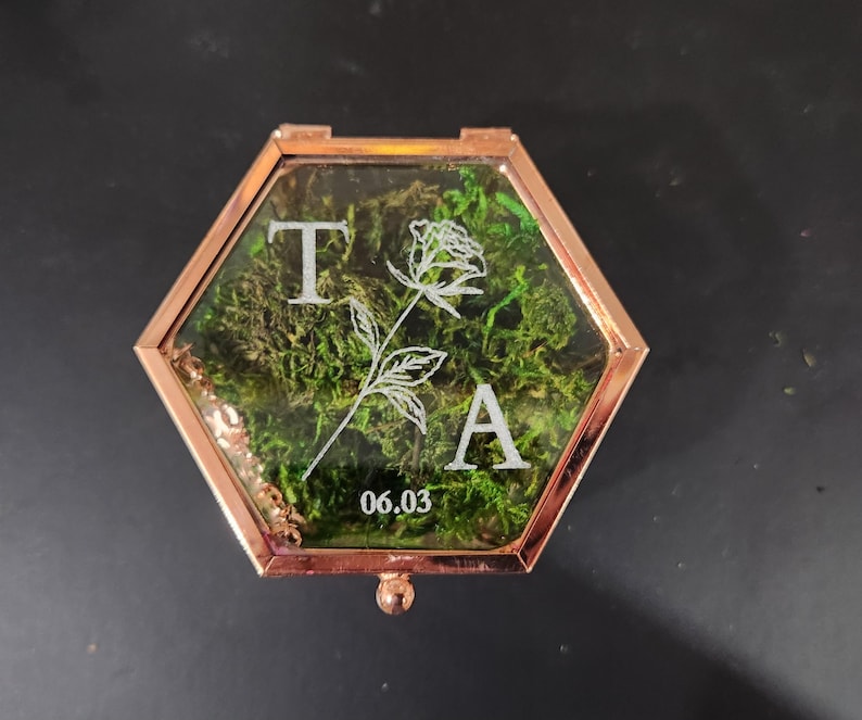 K&ouml;nnte beinhalten: Eine sechseckige Glasschatulle mit einem ros&eacute;goldenen Metallrahmen, gef&uuml;llt mit gr&uuml;nem Moos. Der Glasdeckel ist mit wei&szlig;em Text graviert, darunter die Buchstaben T und A, eine Strichzeichnung einer Rose und das Datum 06.03.