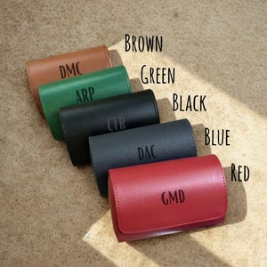 May include: Five rectangular leather cases arranged diagonally in brown, green, black, blue, and red. Each case features a flap closure and is personalised with three-letter monograms: DMC, ARP, CTR, DAC, and GMD, respectively.