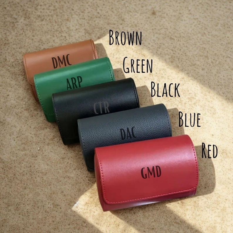 May include: Five rectangular leather cases arranged diagonally in brown, green, black, blue, and red. Each case features a flap closure and is personalised with three-letter monograms: DMC, ARP, CTR, DAC, and GMD, respectively.