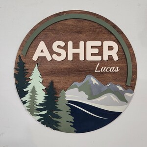 May include: A round wooden sign with the name "ASHER" in white block letters, and the name "Lucas" in script. The sign features a mountain and forest scene with green and blue accents.