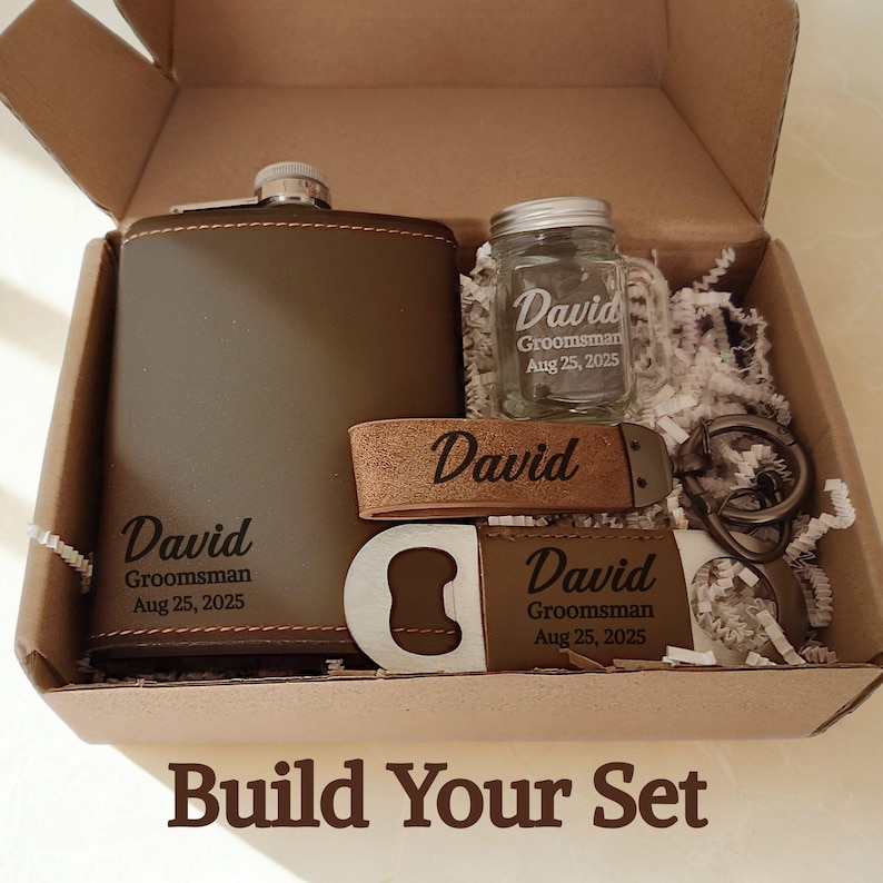 May include: A gift set in a cardboard box, including a brown leather-wrapped hip flask, a small glass jar, a bottle opener, a keyring, and a leather band. Each item is engraved with the name "David" and the date "Aug 25, 2025". The text "Build Your Set" is at the bottom.