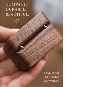 May include: A small, rectangular wooden ring box with a visible grain pattern and gold-toned metal hinges. The text on the box reads: COMPACT. DURABLE. BEAUTIFUL. and TAKE YOUR TREASURES ANYWHERE, accompanied by a small heart symbol.