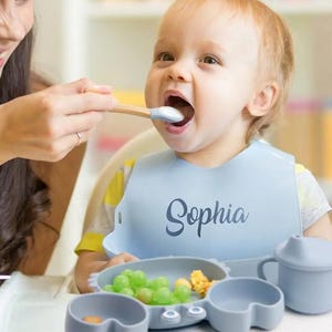 K&ouml;nnte beinhalten: Ein Baby tr&auml;gt eine hellblaue L&auml;tzchen mit dem Namen "Sophia" in Schreibschrift. Das Baby wird mit einem L&ouml;ffel gef&uuml;ttert. Ein grauer Baby-Teller aus Silikon mit Essen und ein Trinkbecher befinden sich vor dem Baby.