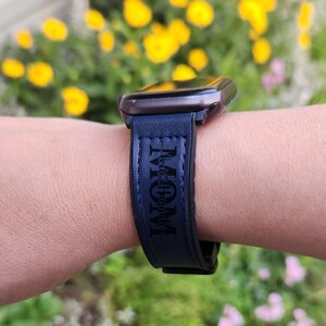 Pu&ograve; includere: Un cinturino per smartwatch blu scuro con la parola "MOM" stampata verticalmente in un carattere stilizzato. Il cinturino &egrave; attaccato a uno smartwatch nero. Lo sfondo &egrave; sfocato, con fiori gialli visibili.