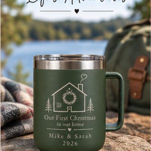 May include: A matte olive green stainless steel mug with a silver rim and handle. The mug features an engraved design of a house with a wreath, pine trees, and the text "Our First Christmas in our home", "Mike & Sarah", and the year "2026".